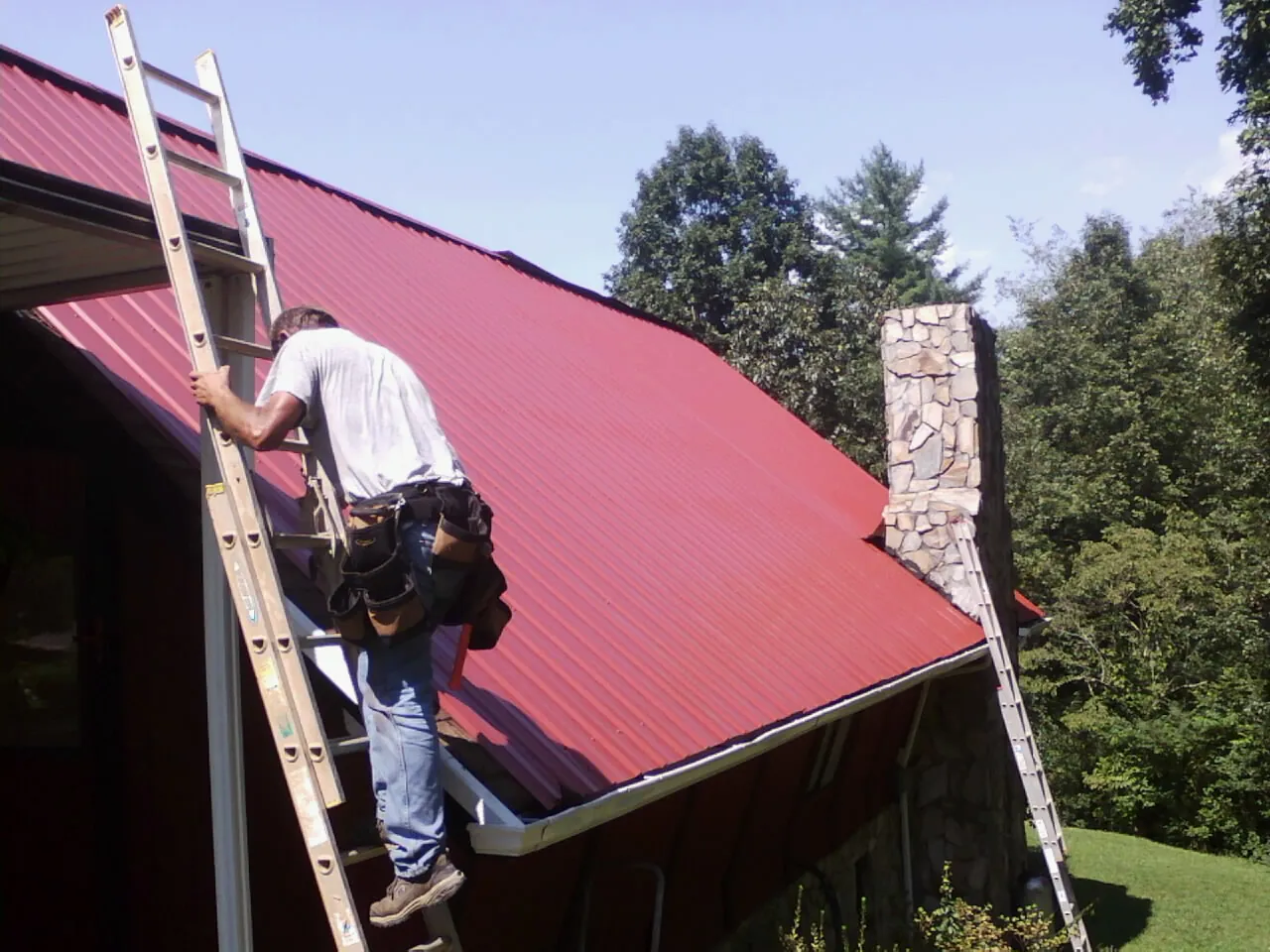 Roofer on a ladder beside a red metal roof and stone chimney — Soffit & Fascia Repair by Roofing Brothers in Divernon